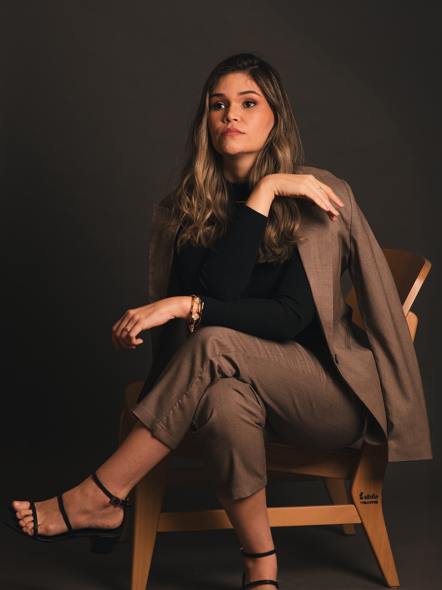 Lays Maciel sentada em uma cadeira de madeira, de gola alta preta e blazer taupe nos ombros, sob luz direcional.