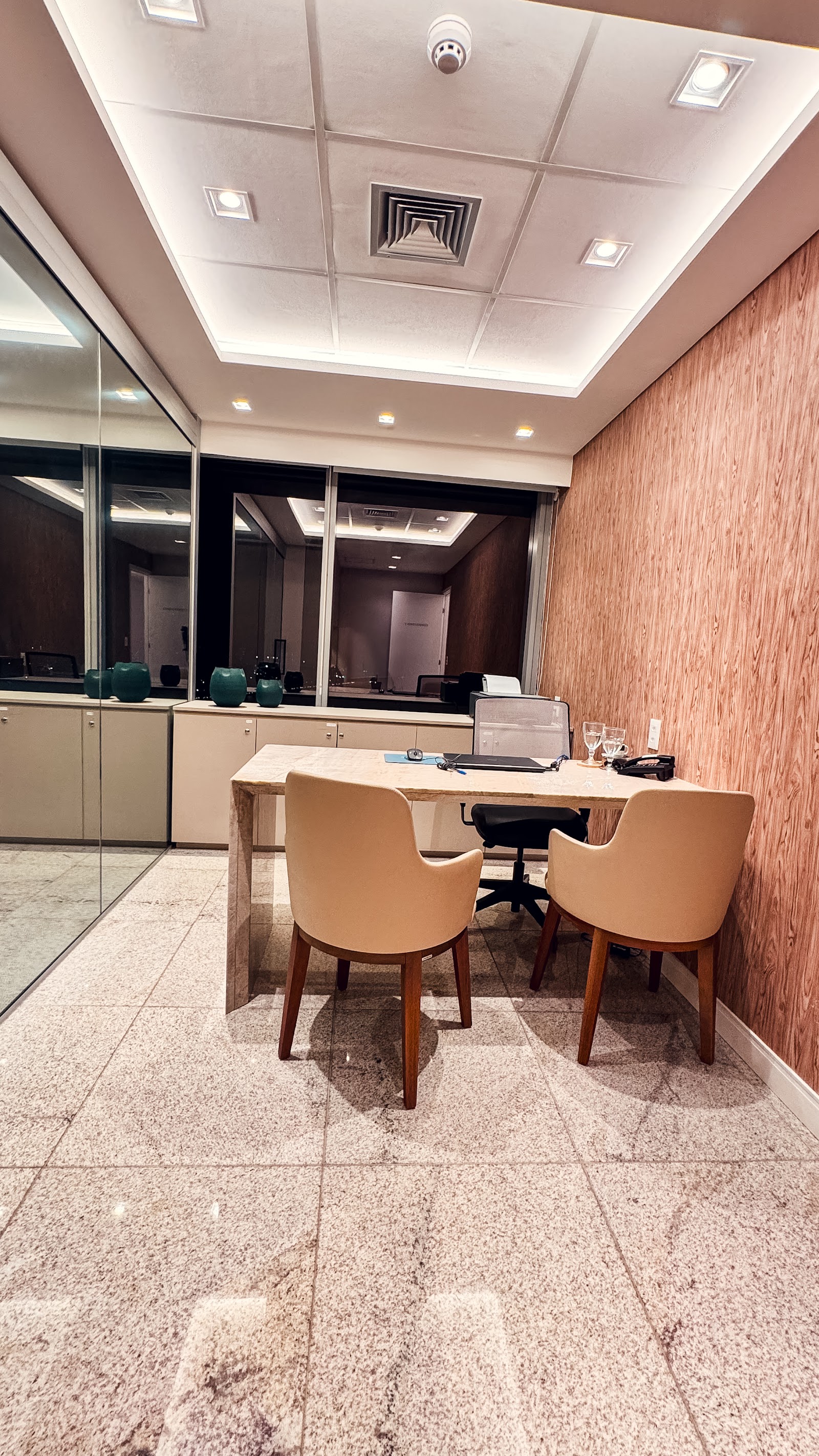 A sala do consultório: parede de ripas verticais de madeira acesa por spots, mesa de mármore claro e cadeiras bege.