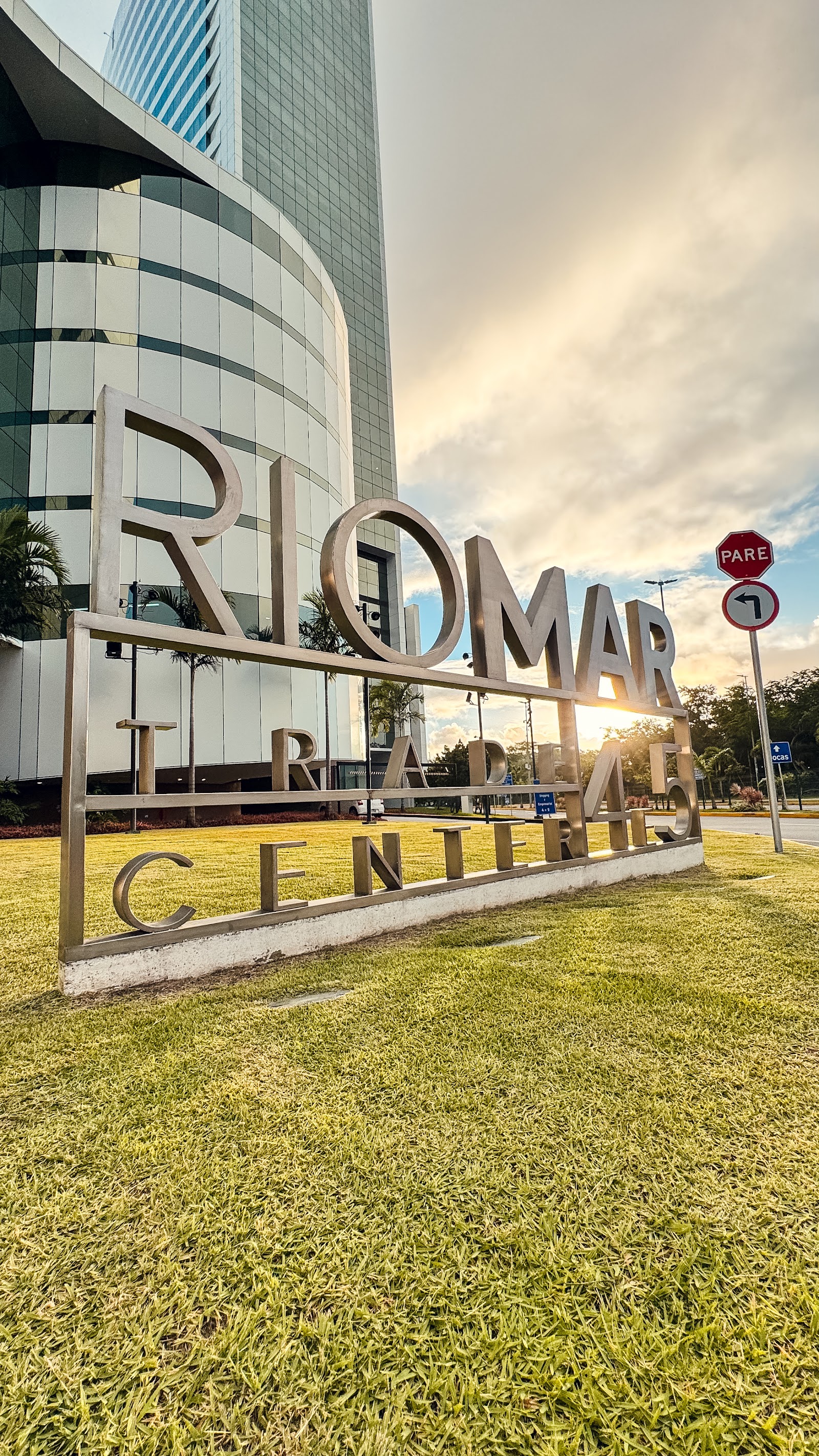 O letreiro RioMar Trade Center em letras de metal sobre a grama, com o sol poente por trás.