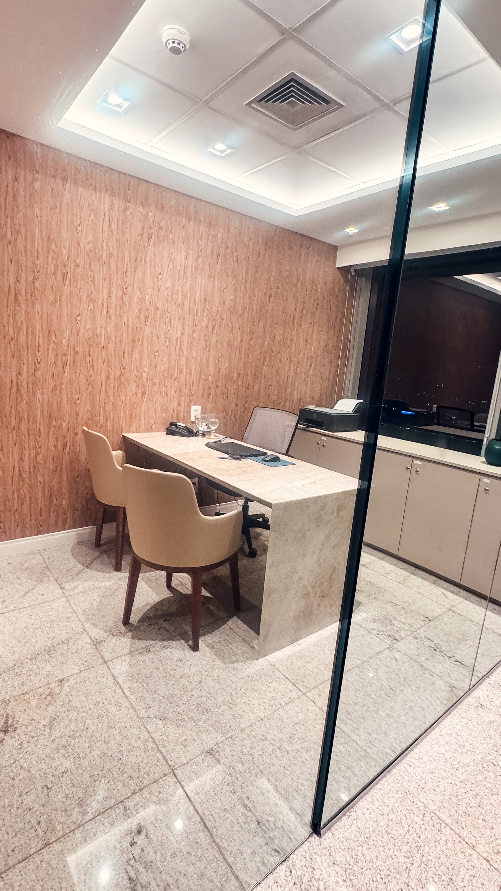 A sala vista do corredor, com o ripado à esquerda, a mesa de mármore ao centro e a janela da cidade apagada atrás.